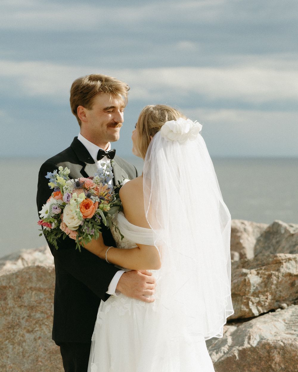 A romantic seaside wedding scene with a couple embracing on a rocky beach under an overcast sky.