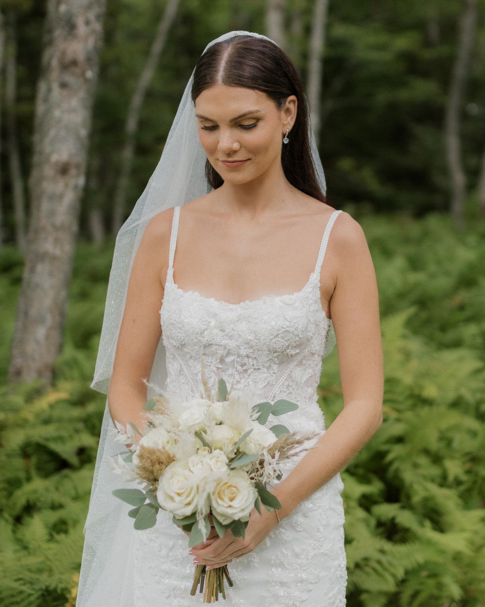 A bride in a white lace wedding dress holds a beautiful bouquet of white roses and greenery in a forest setting.