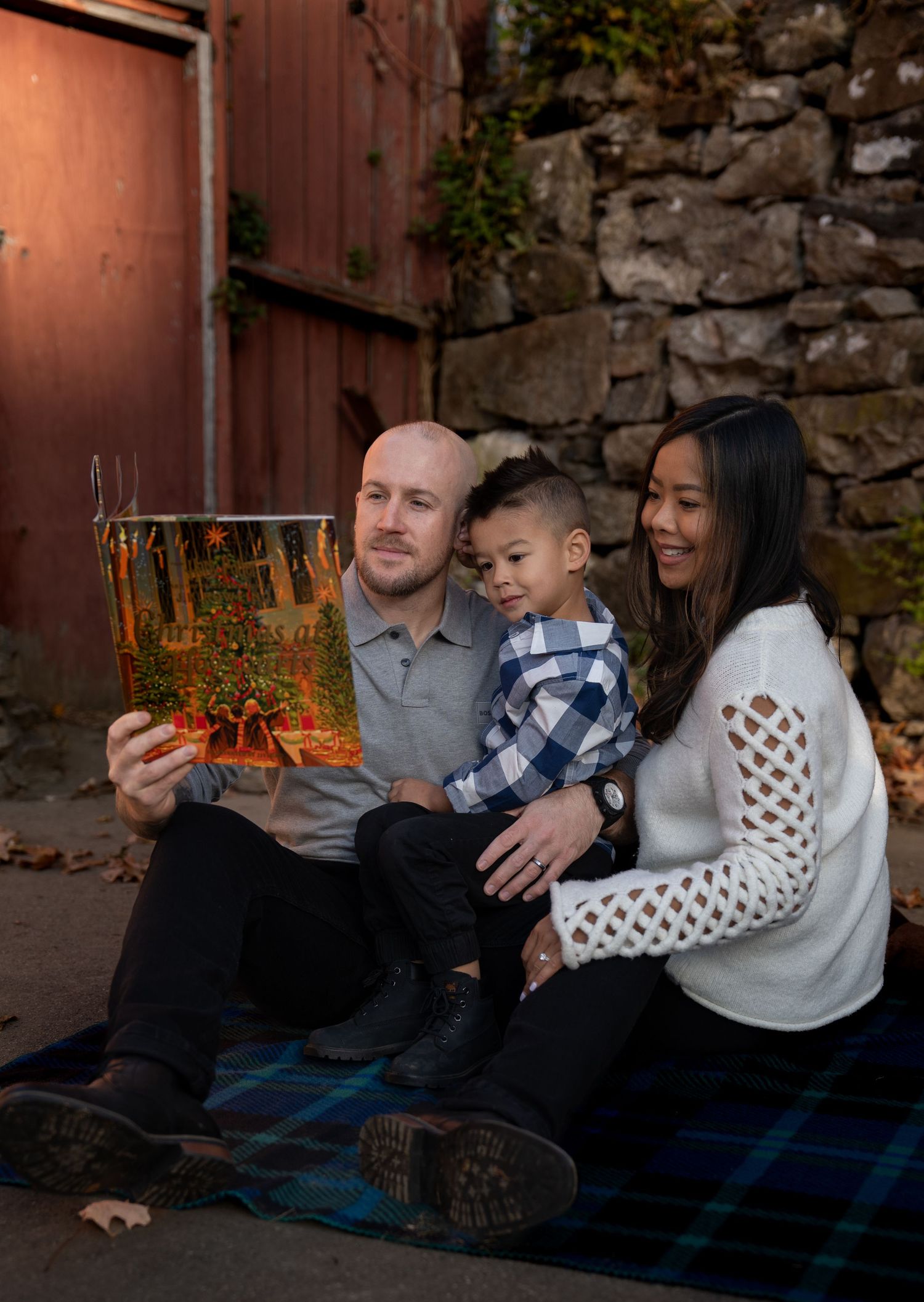 Family sitting together on a plaid blanket against a rustic stone wall.