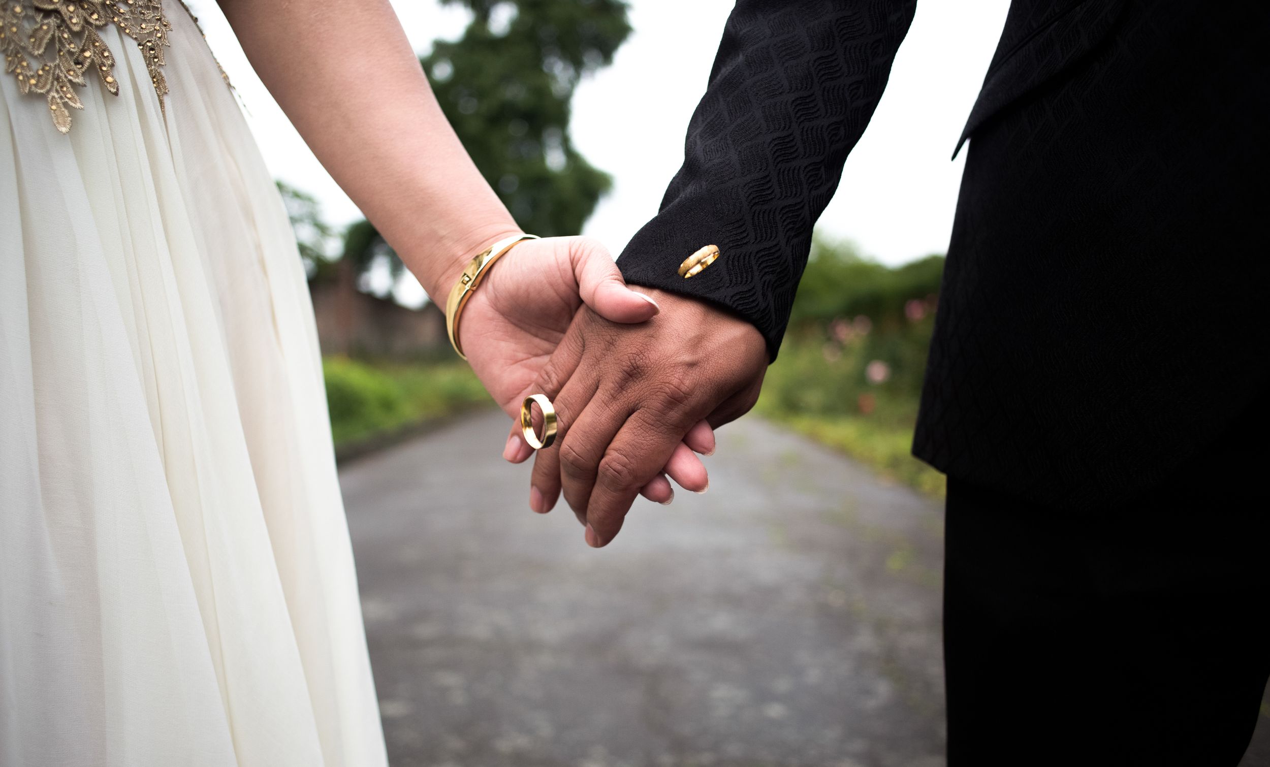 Close-up of newlywed couple holding hands on a path, showing white dress and black suit details.
