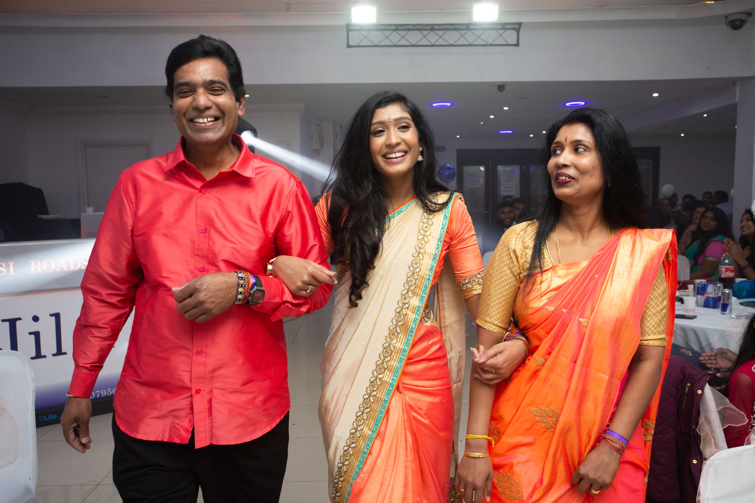 Three people in festive attire smile together at an indoor event, wearing bright red and orange traditional clothing.