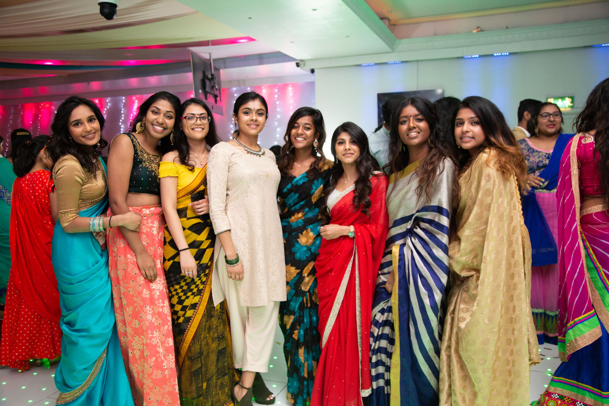A colorful celebration with people wearing traditional Indian saris and formal attire at an indoor party venue.