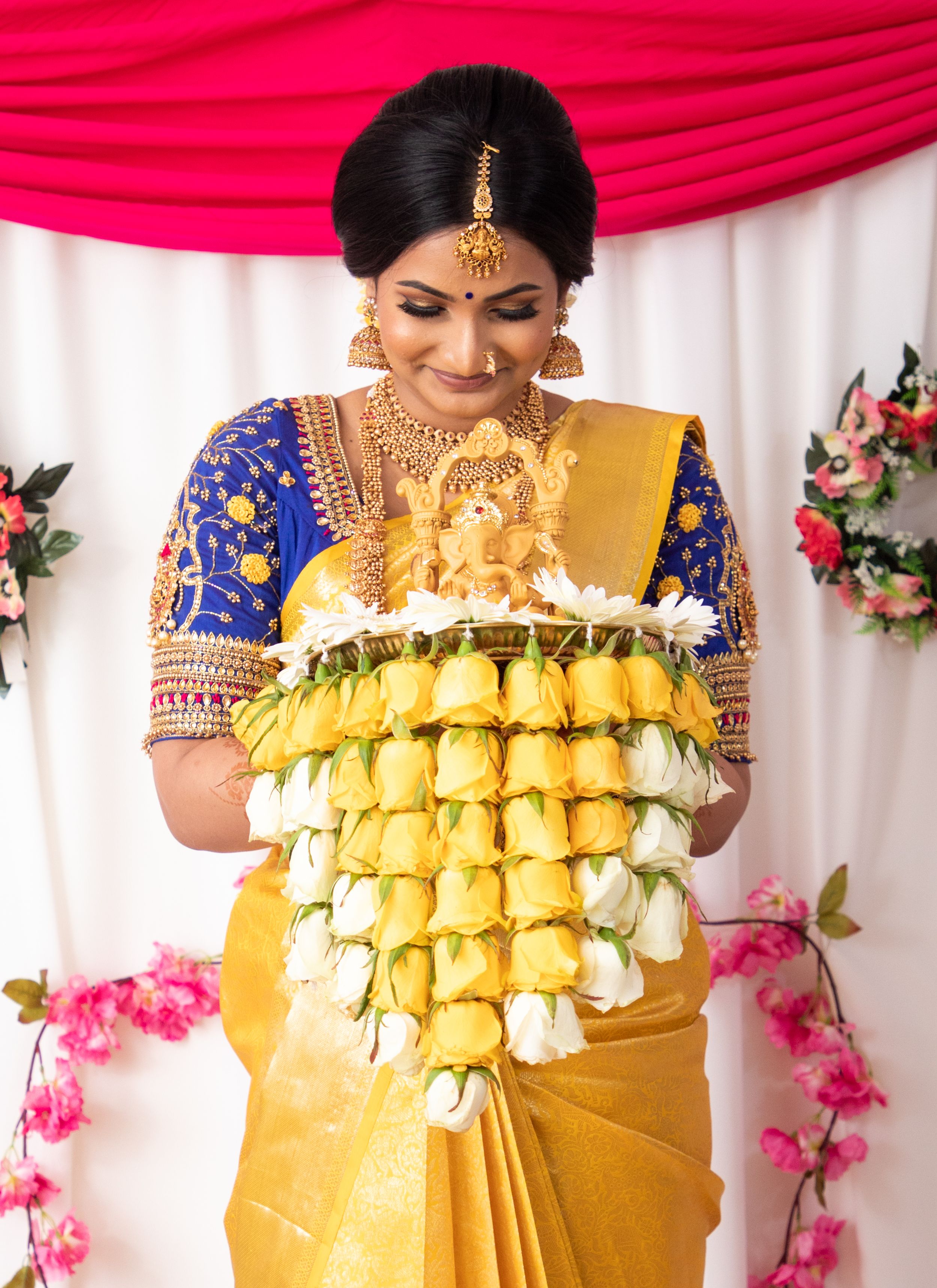 Woman in traditional Indian attire with elaborate jewellery holds a decorative floral garland against backdrop of pink flowers.