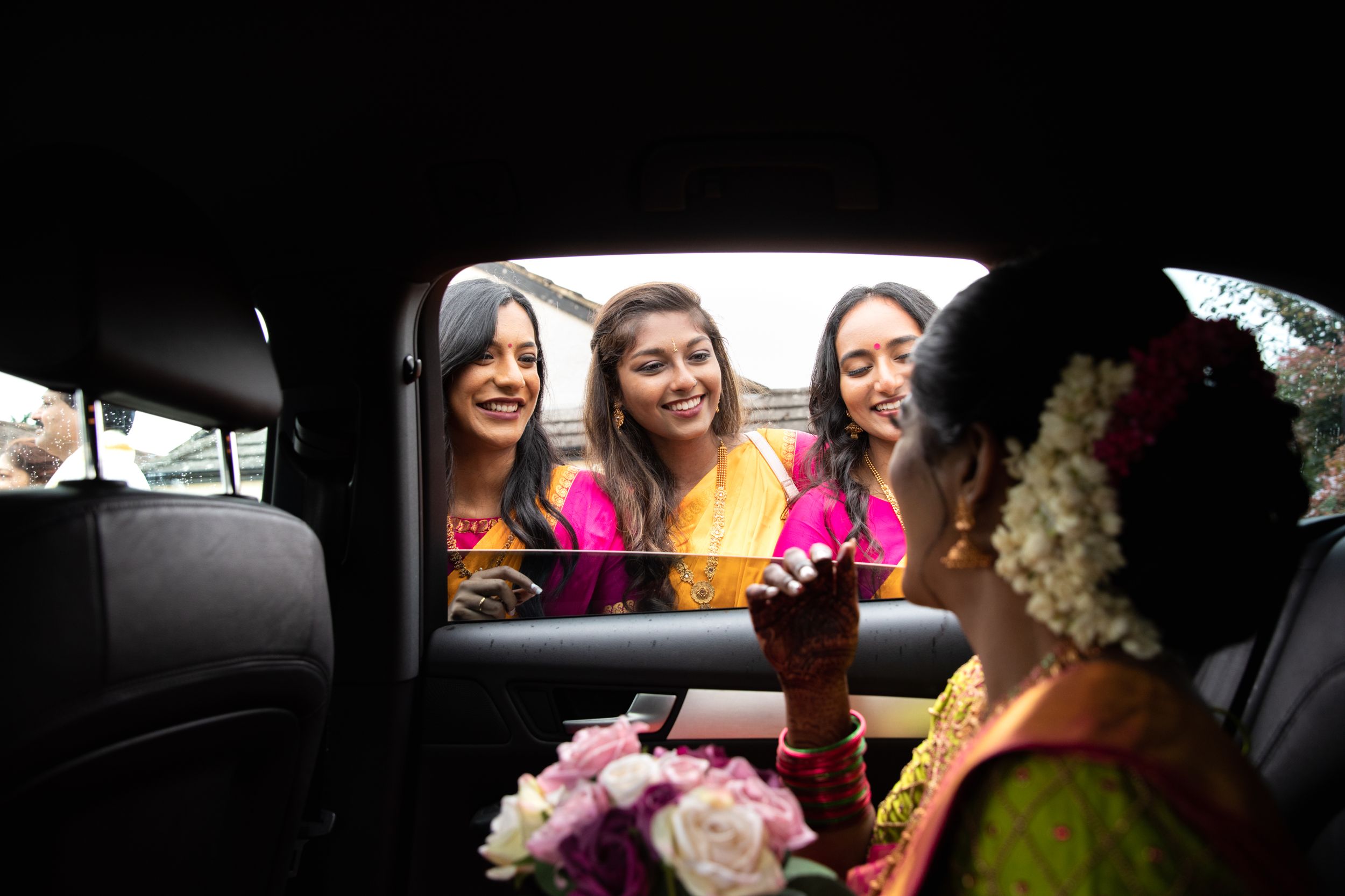 Three people peer through a car window at someone inside while smiling and laughing.