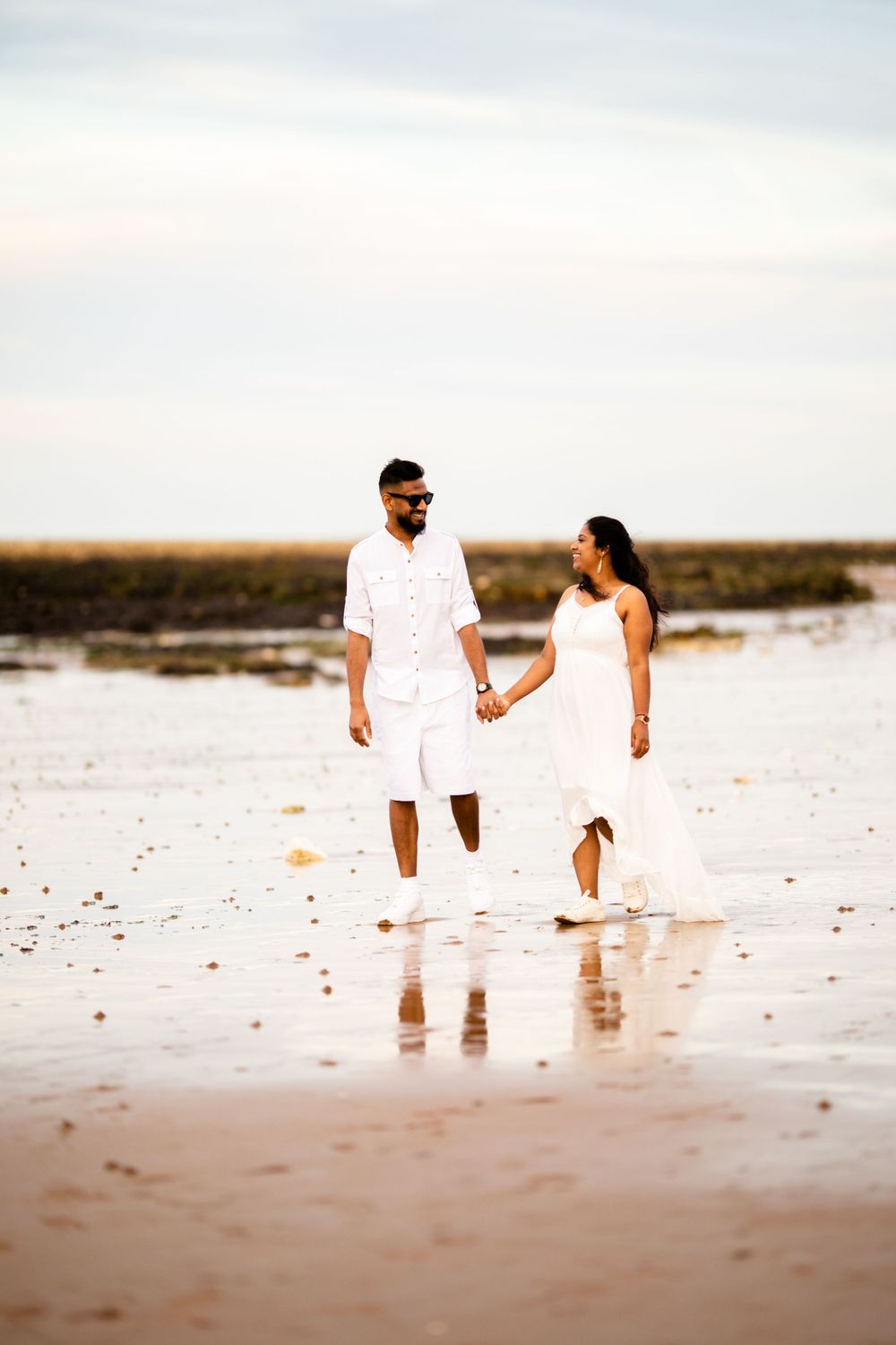 A couple dressed in white holds hands while walking along a serene beach at sunset, their reflections visible in the wet sand.