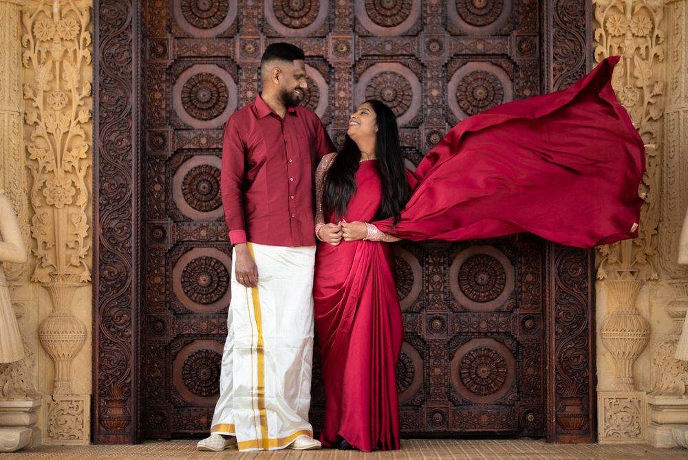 A couple in matching red traditional Indian outfits pose against an ornate carved door with flowing fabric in motion.