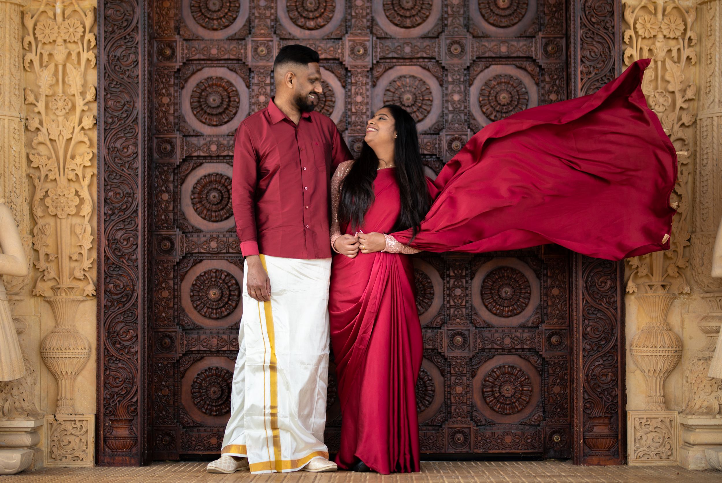 A couple in matching red traditional Indian outfits pose against an ornate carved door with flowing fabric in motion.