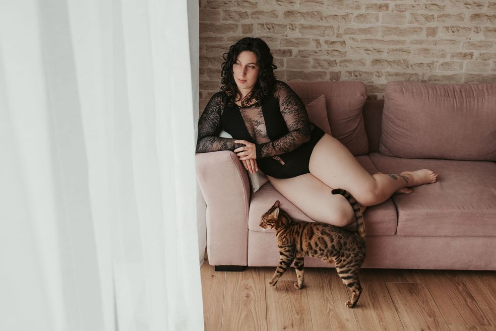 Woman in black dress lounges on pink sofa in brick-walled room with natural lighting.