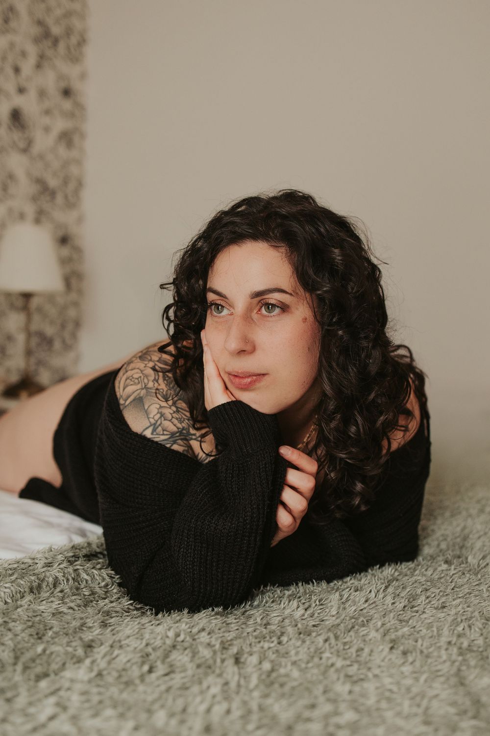 A person with dark curly hair wearing a black sweater lies on a gray carpet in a cozy indoor setting.