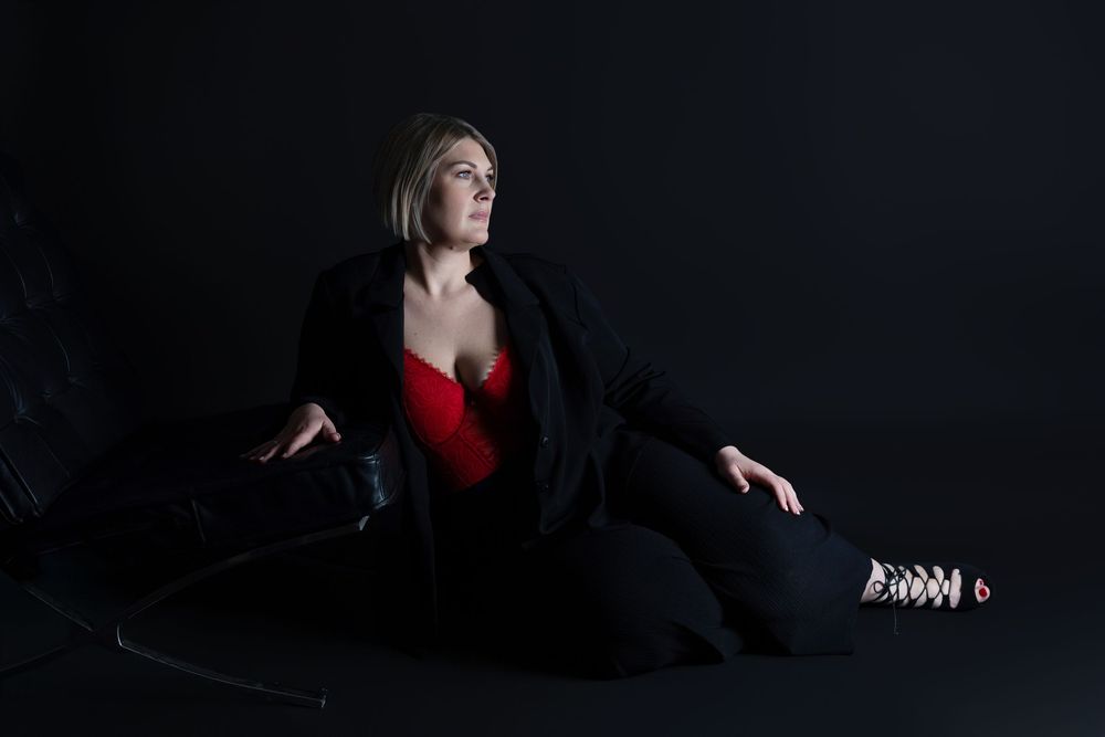 Dramatic studio portrait with figure in red dress and black blazer posed against dark background.
