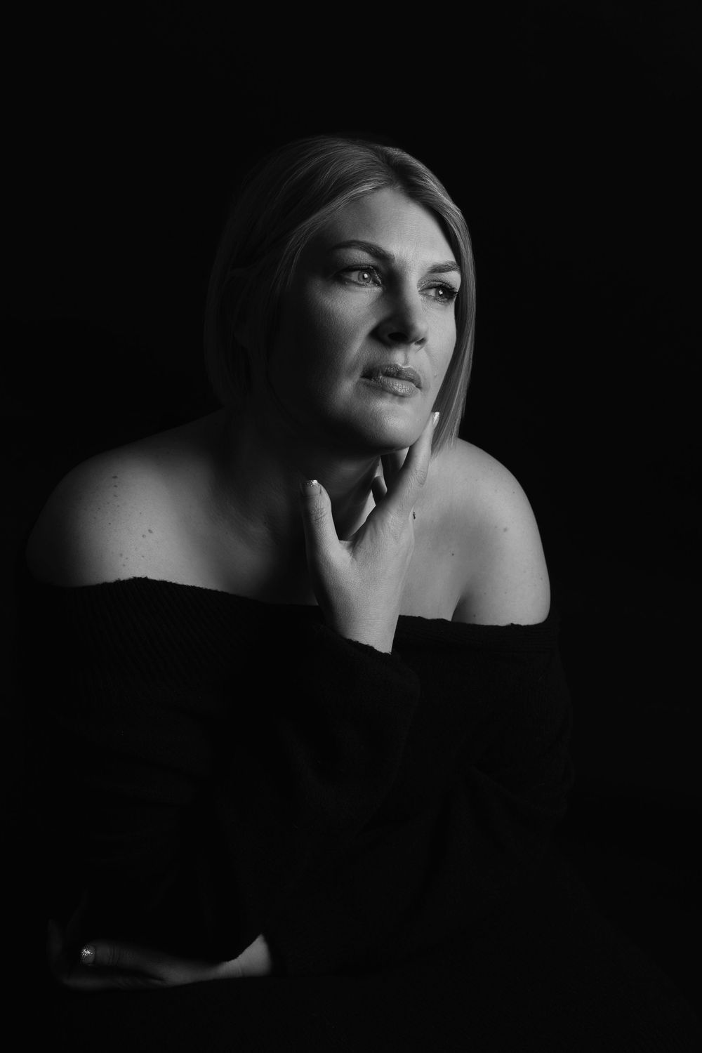 A dramatic black and white portrait of a woman in an off-shoulder pose with soft lighting and contemplative expression.