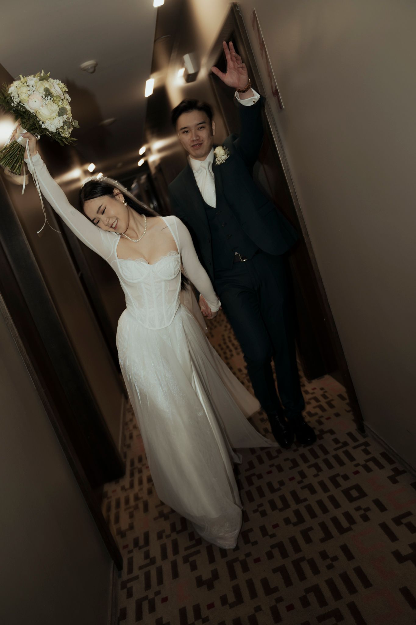 Bride and groom celebrating in hallway with raised bouquet and arms.