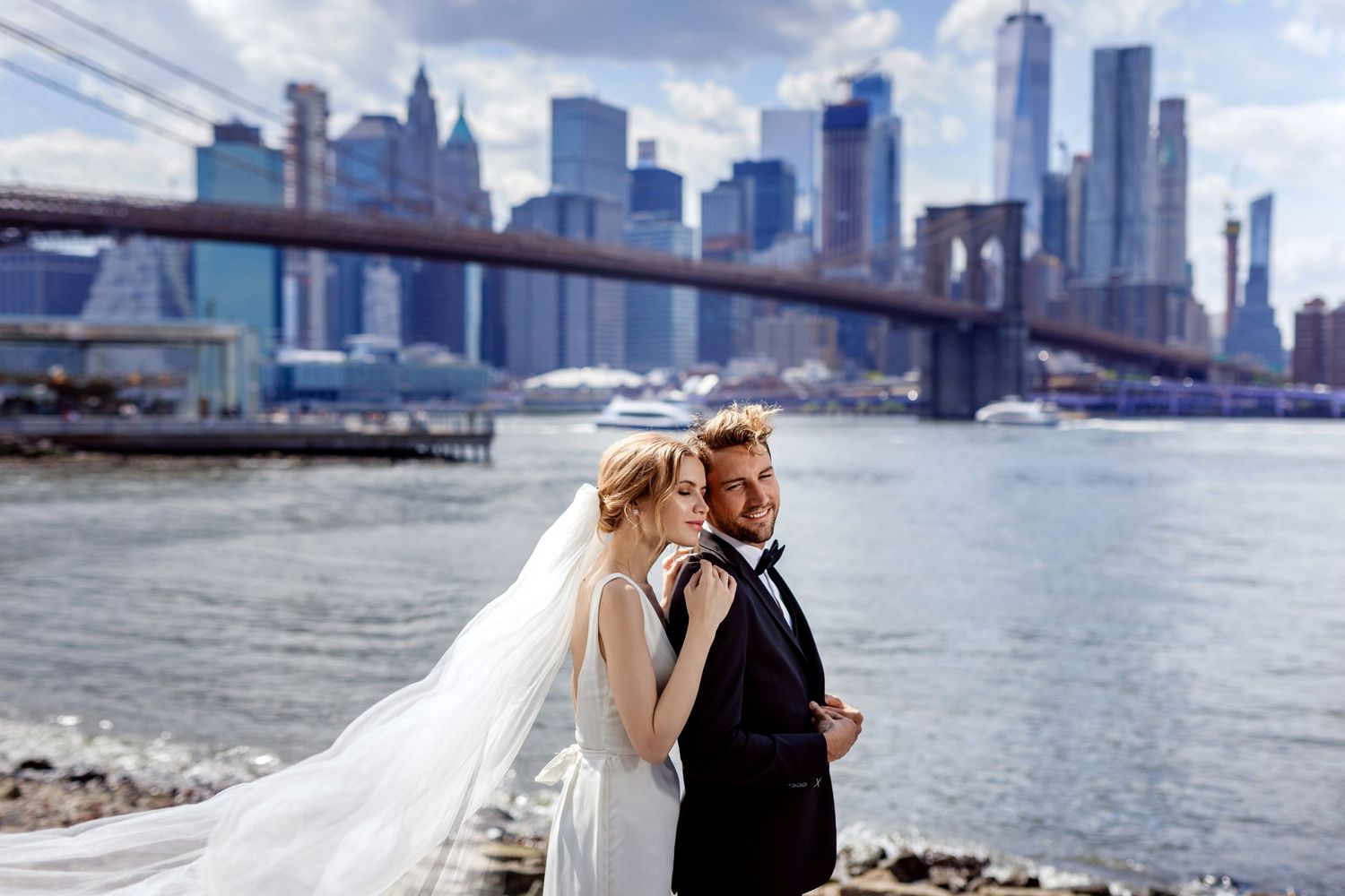 A couple in wedding attire embraces near the Brooklyn Bridge with the Manhattan skyline in the background.