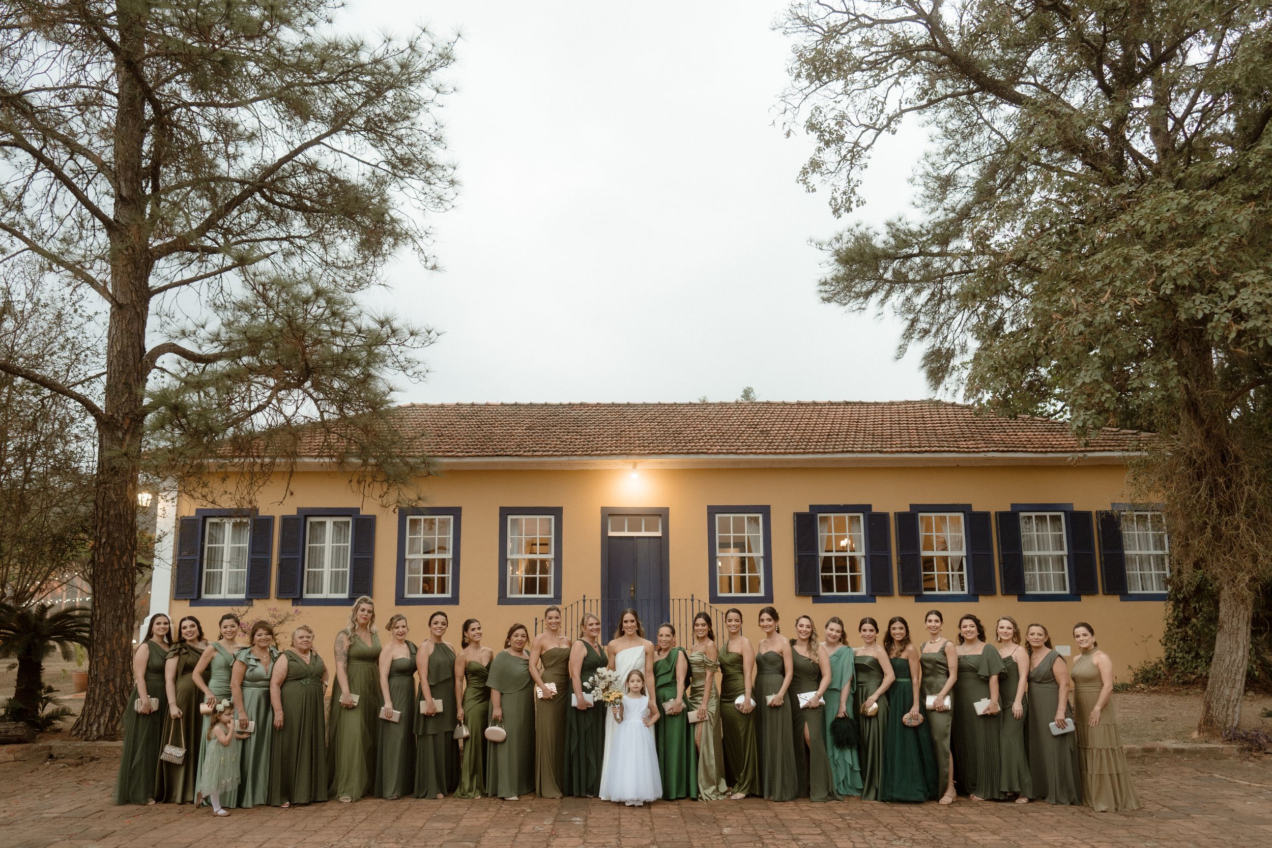 A wedding party in green dresses and gray suits poses in front of a historic colonial-style building.