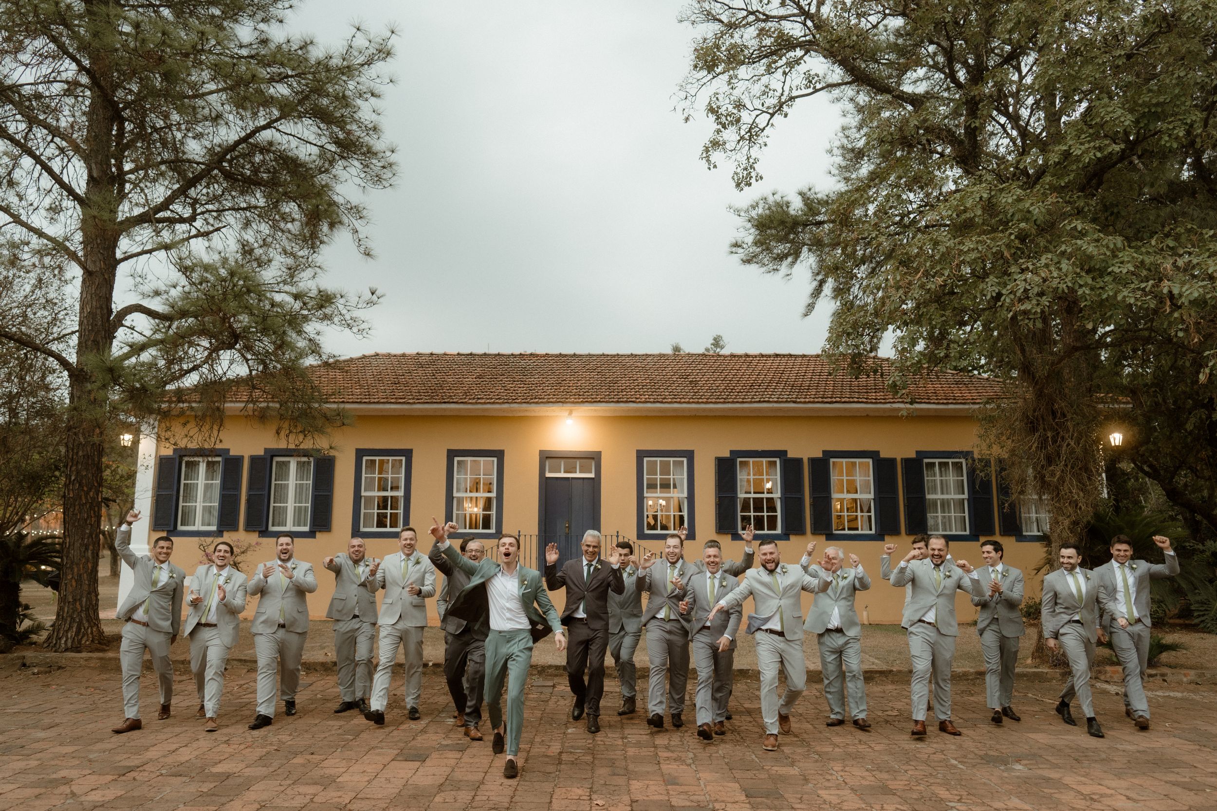 Groomsmen in light suits line up in front of a historic building with black shutters.