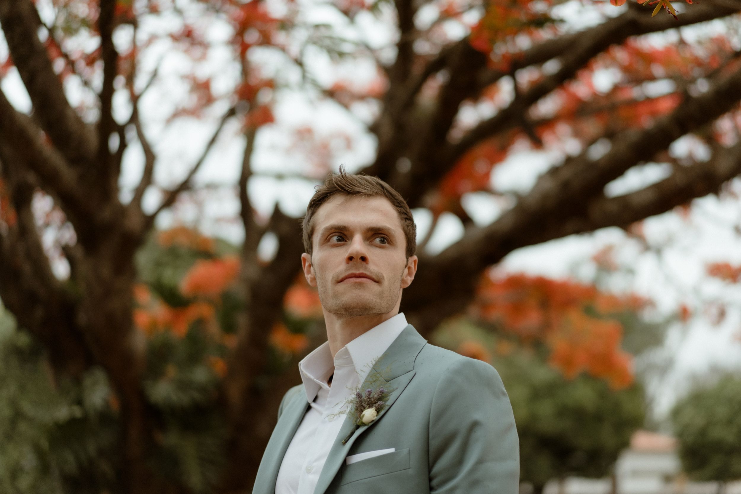 A formal portrait of a groom in a light gray suit against autumn foliage.