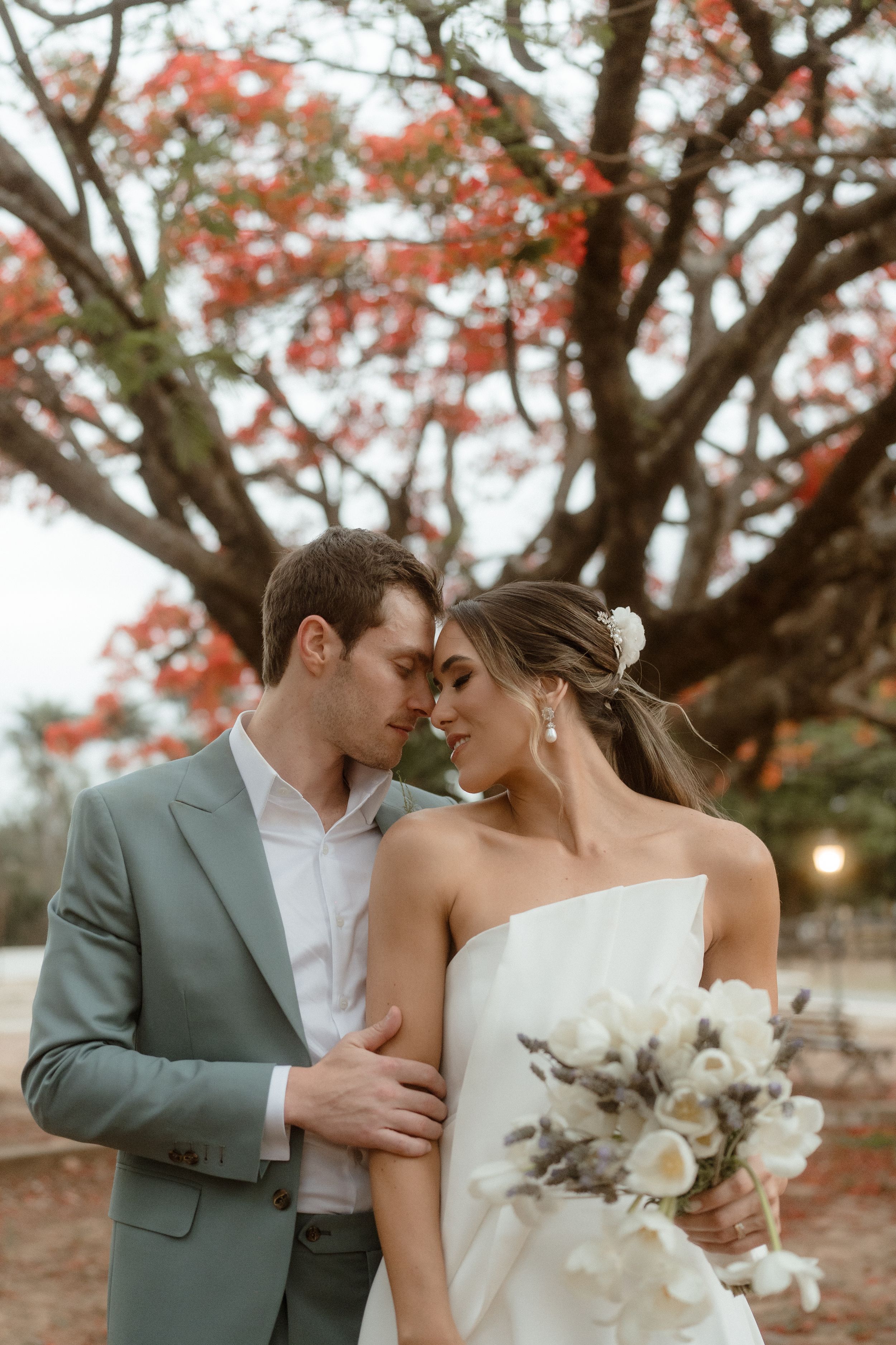 A tender moment between a couple under a blooming tree with the bride holding a white bouquet.