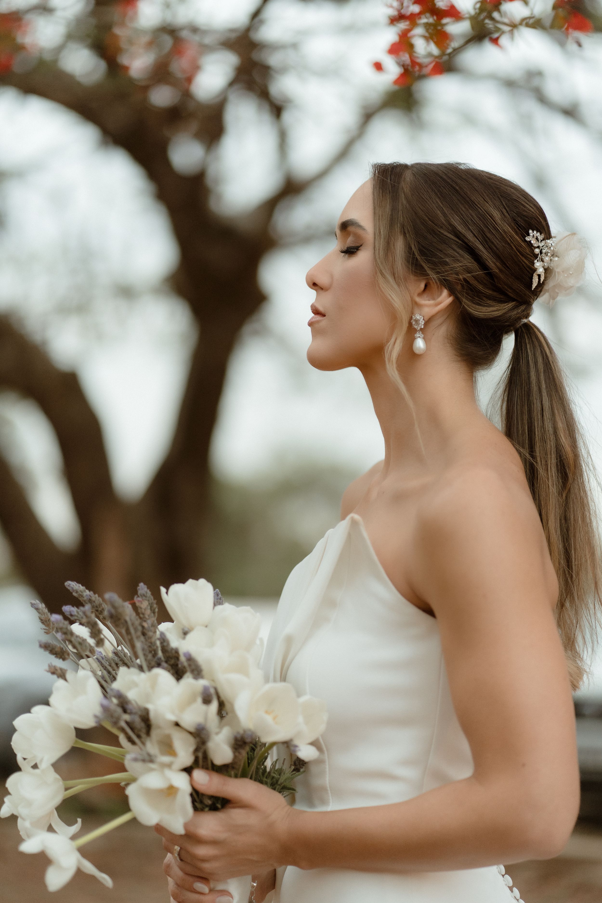 An elegant side profile of a bride with a ponytail holding a white bouquet against autumn trees.