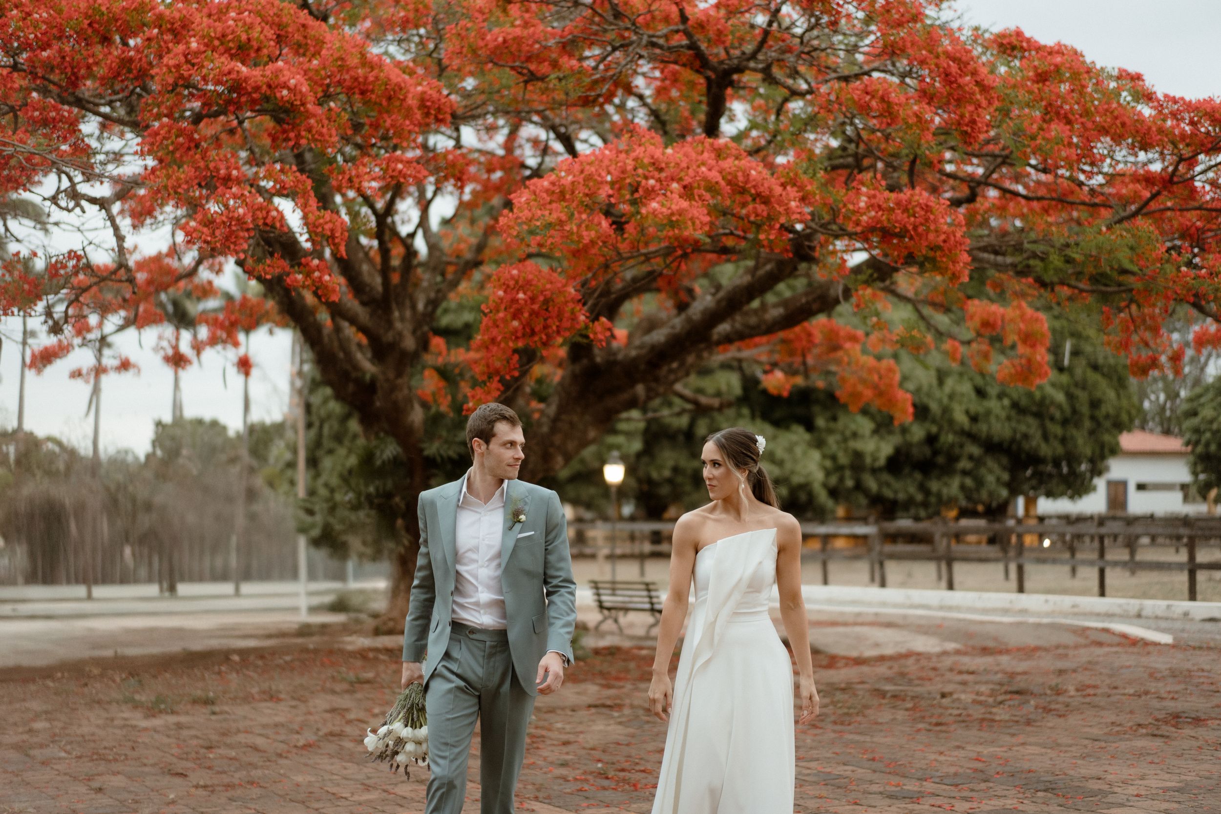 A couple walks hand in hand under vibrant red autumn trees in their wedding attire.