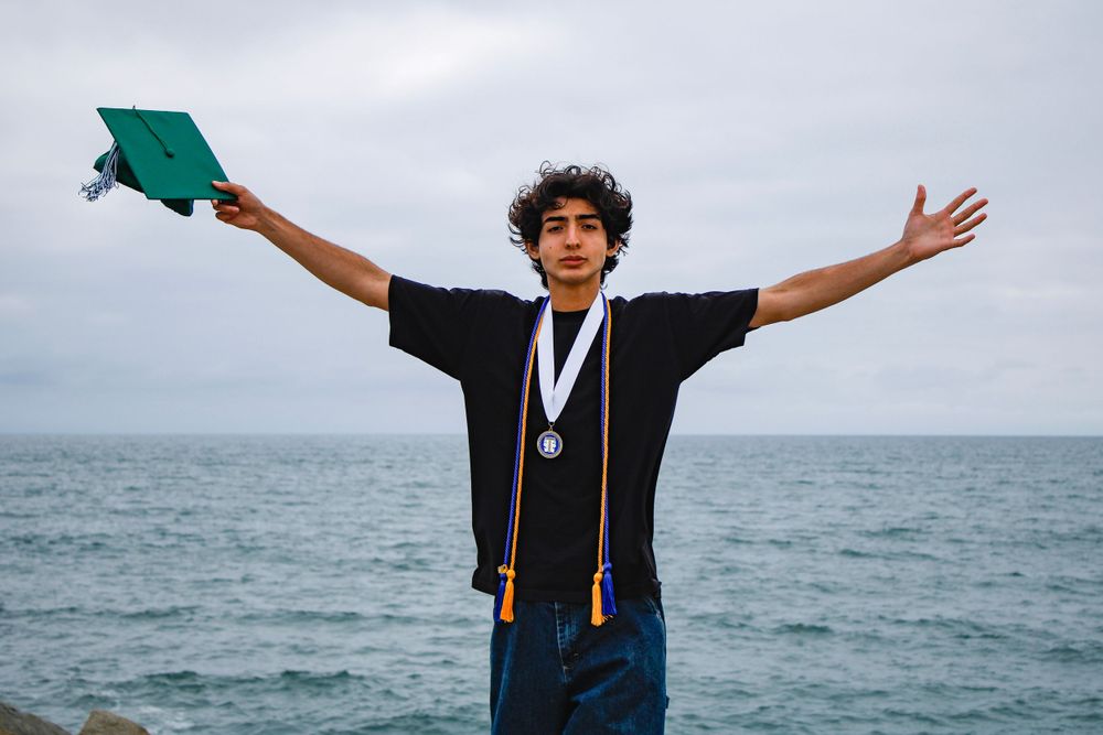 Graduate in emerald green cap and gown celebrating against cloudy ocean backdrop.