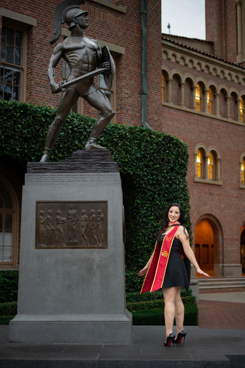 Graduate in black dress poses near historic brick building with statues.