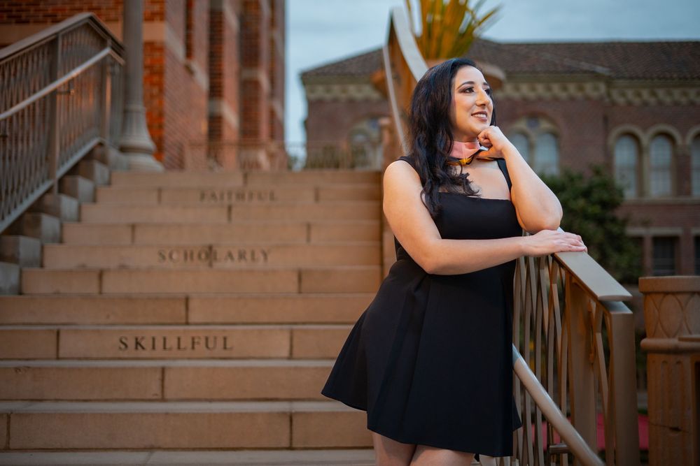 Graduate in black dress poses on steps of brick building.