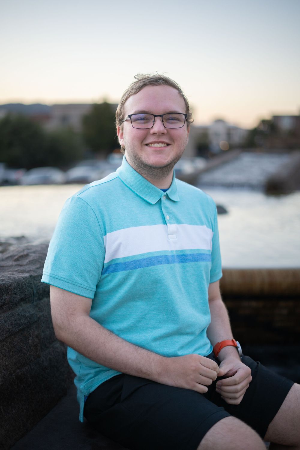 A person in a turquoise polo shirt sits near a waterfront at sunset.