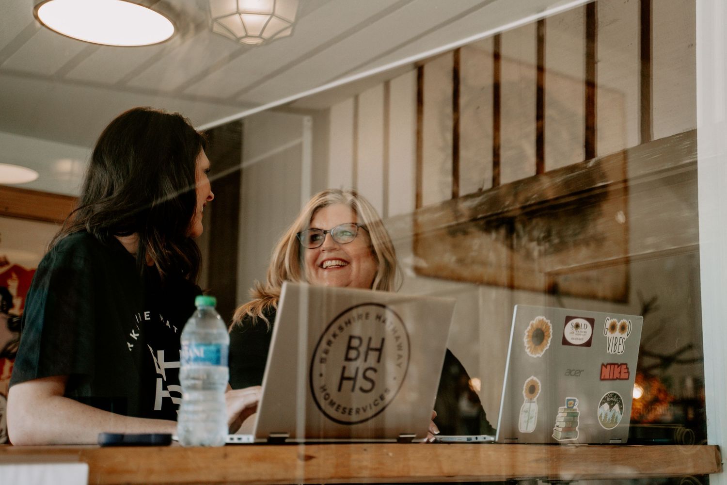 Two people share a laugh while working on laptops in a modern coffee shop.
