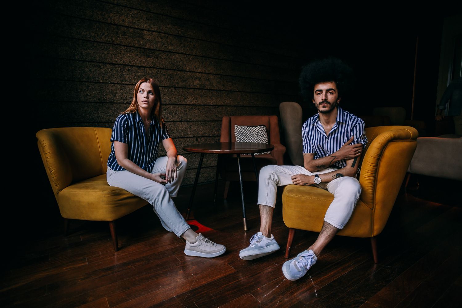 Two people sit in stylish yellow armchairs against a dark wooden wall in a moody, intimate setting.