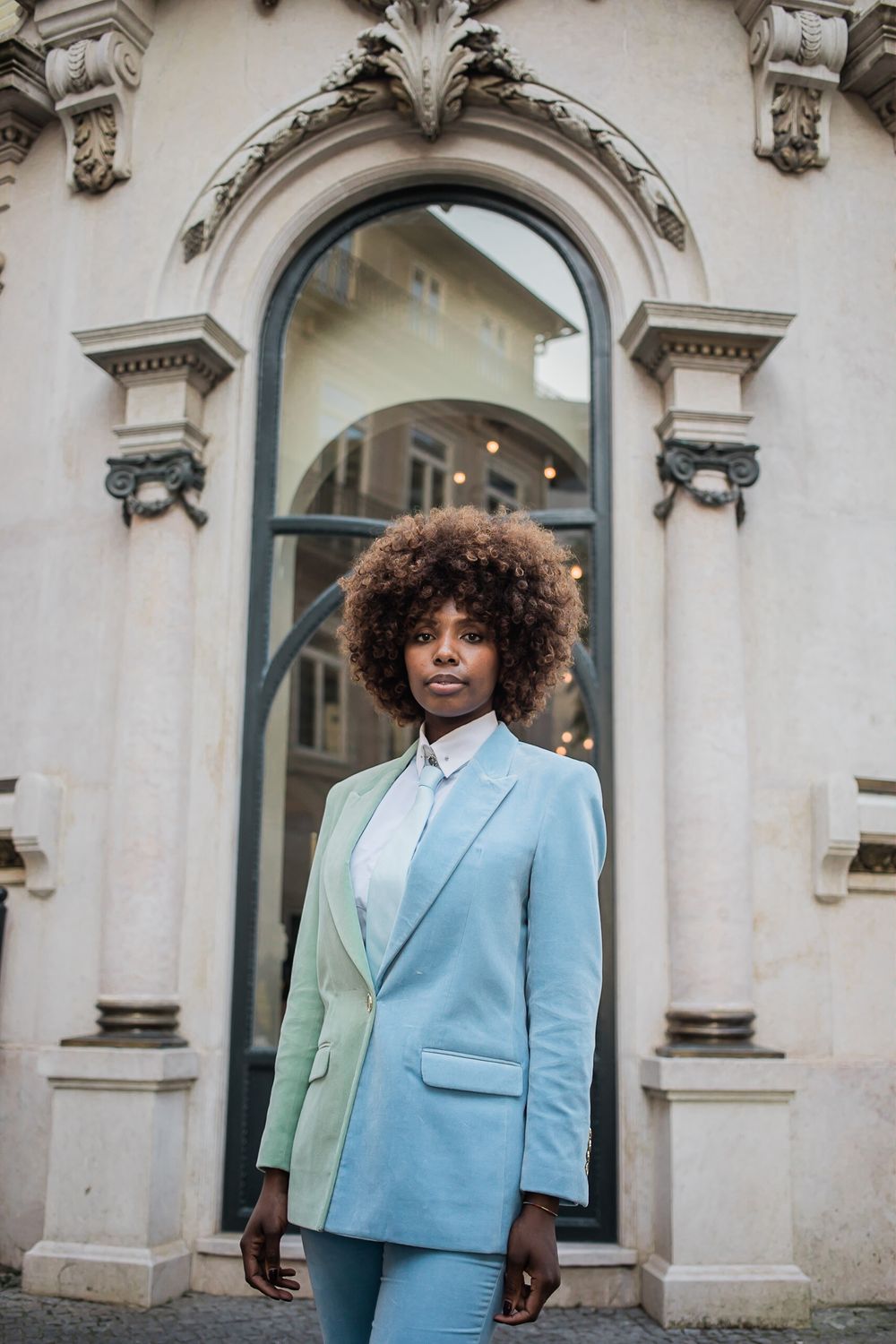 A person wearing a stylish light blue suit poses in front of an ornate architectural doorway.