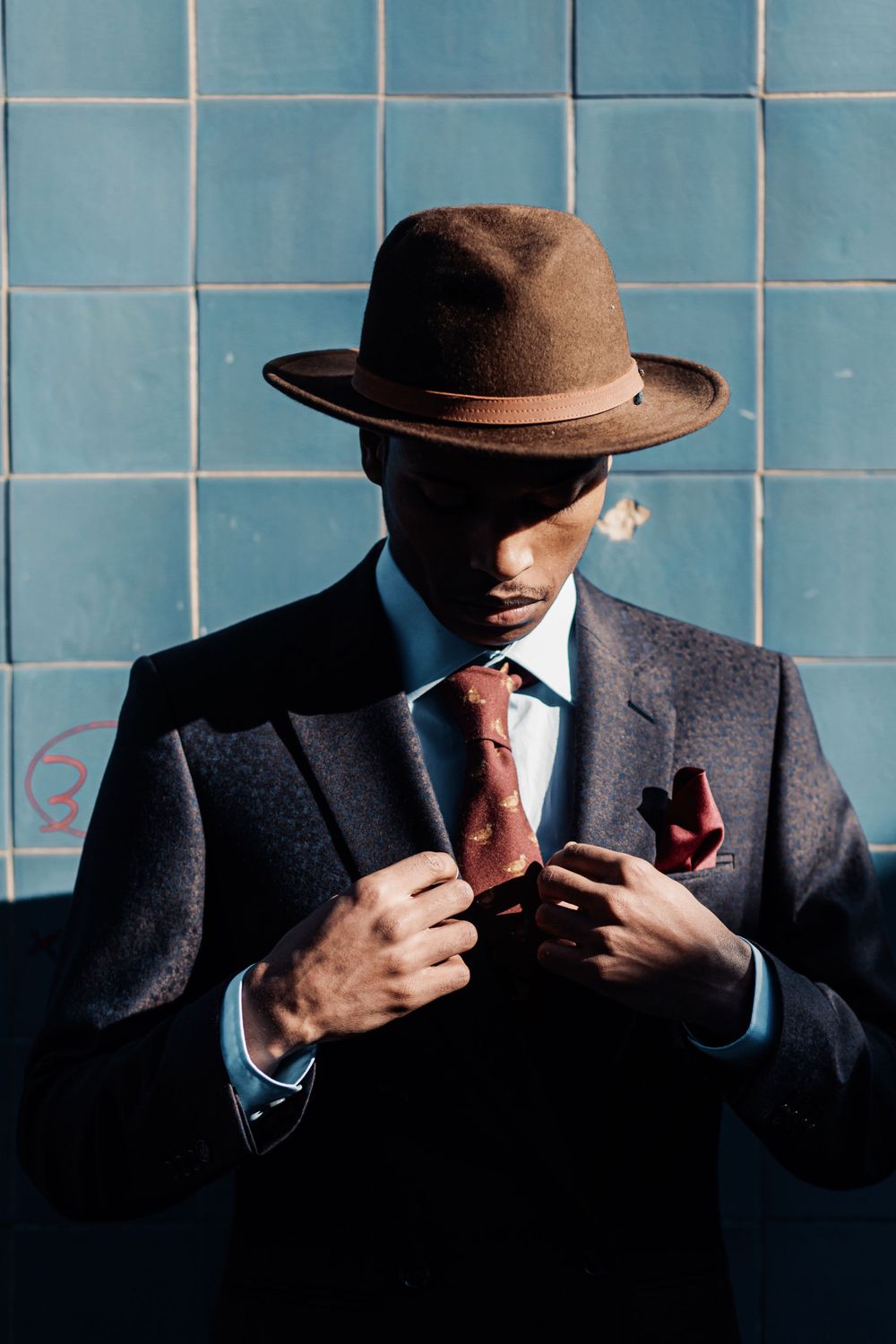 A person in a brown fedora and dark suit adjusts their red tie against a blue tiled wall.