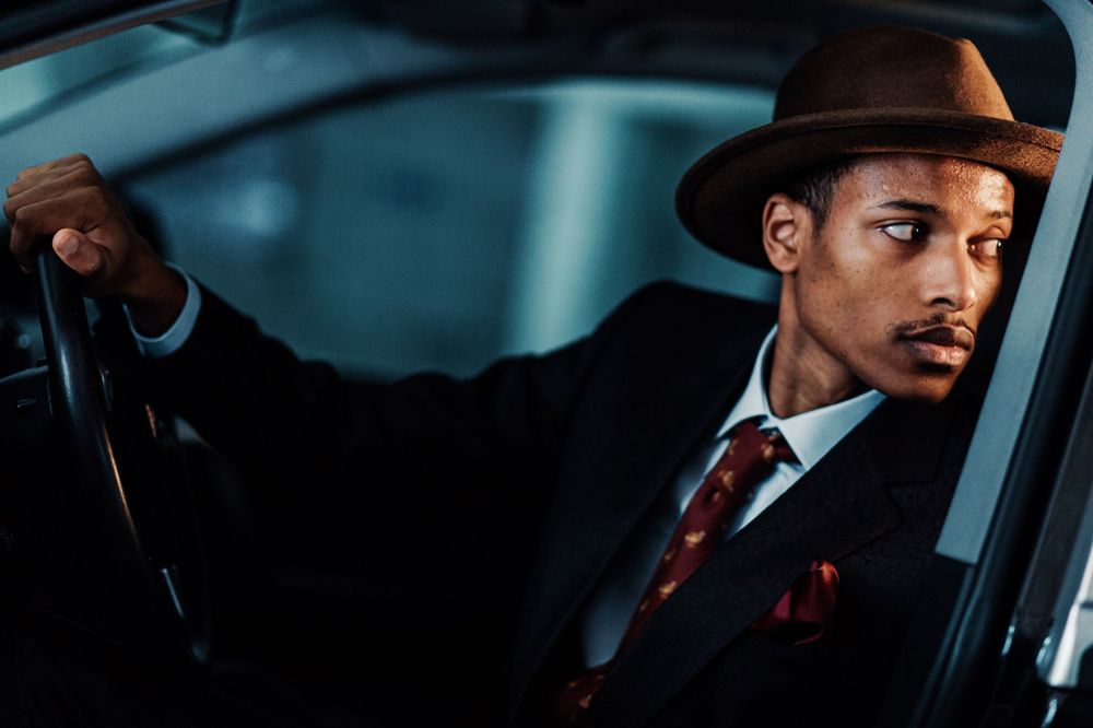 A stylishly dressed person in a brown fedora and red tie sits at the steering wheel of a car in moody lighting.