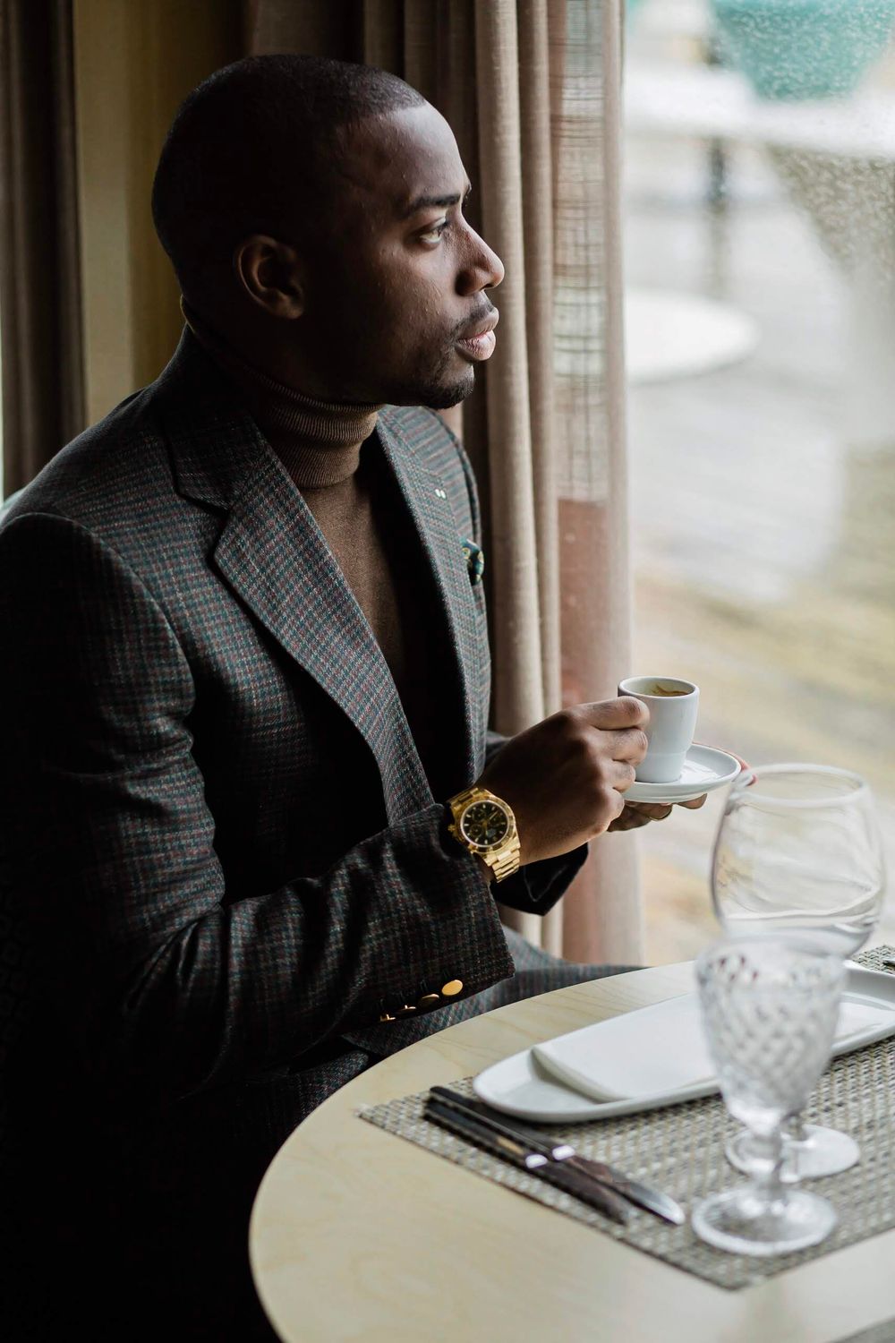 A person in a gray blazer sits at a restaurant table by a window, holding a coffee cup while wearing a gold watch.
