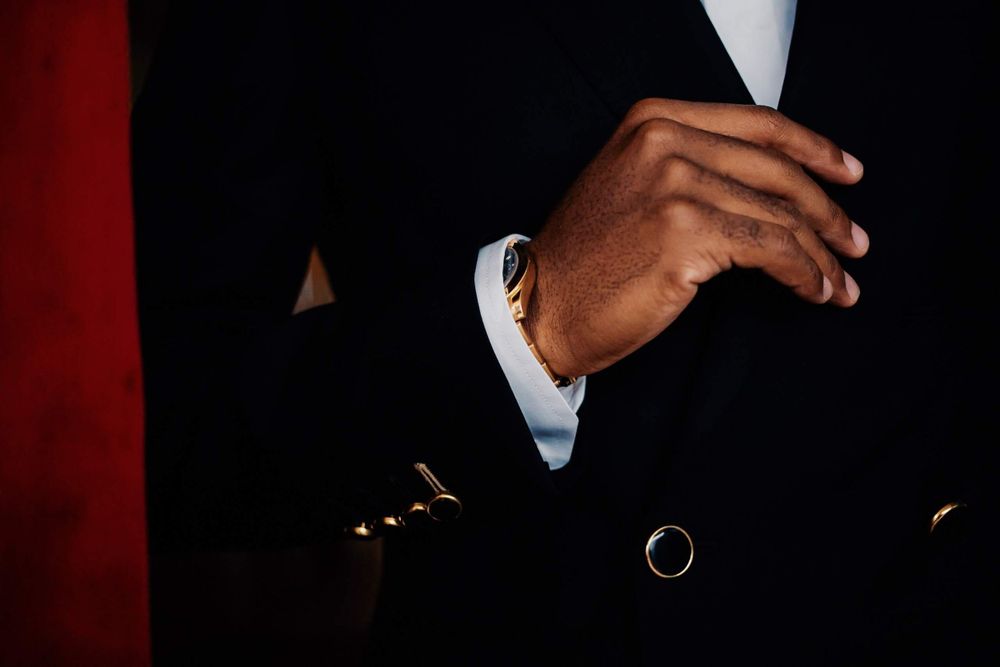 Close up detail of a black formal suit jacket with a white shirt cuff visible against a dark red background.