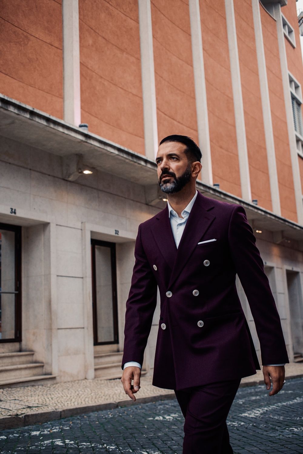A person wearing a stylish double-breasted purple suit walks confidently past an orange building with white columns.