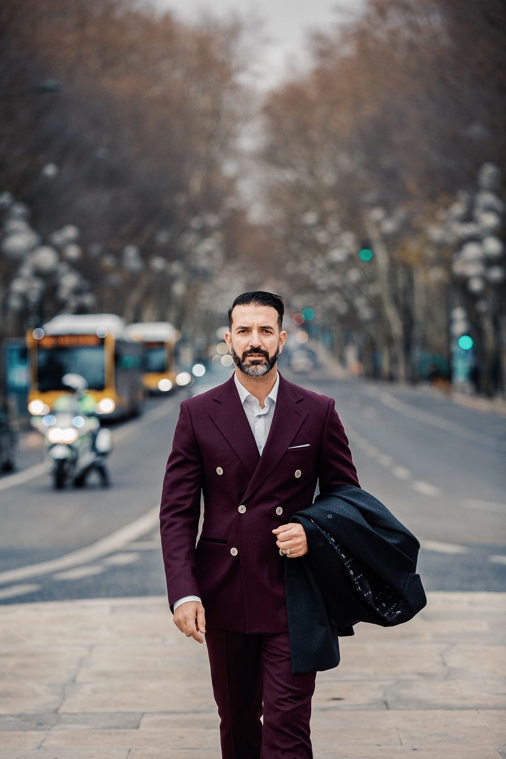 A well-dressed person in a burgundy suit walks down a city street on an autumn day with traffic in the background.