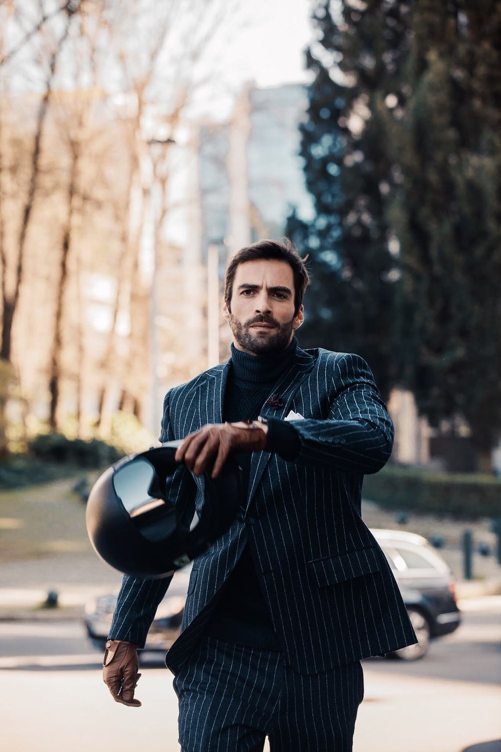 A person in a pinstriped suit carries a motorcycle helmet while walking outdoors on an autumn day.