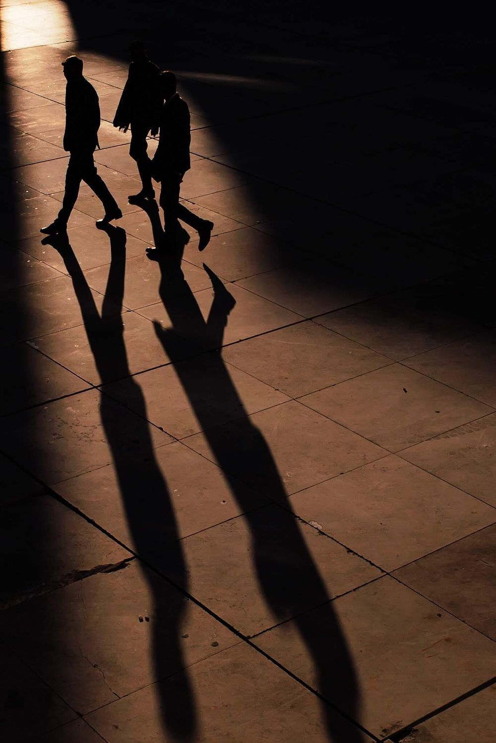 Two elongated shadows cast on a tiled floor from people walking in dramatic lighting.