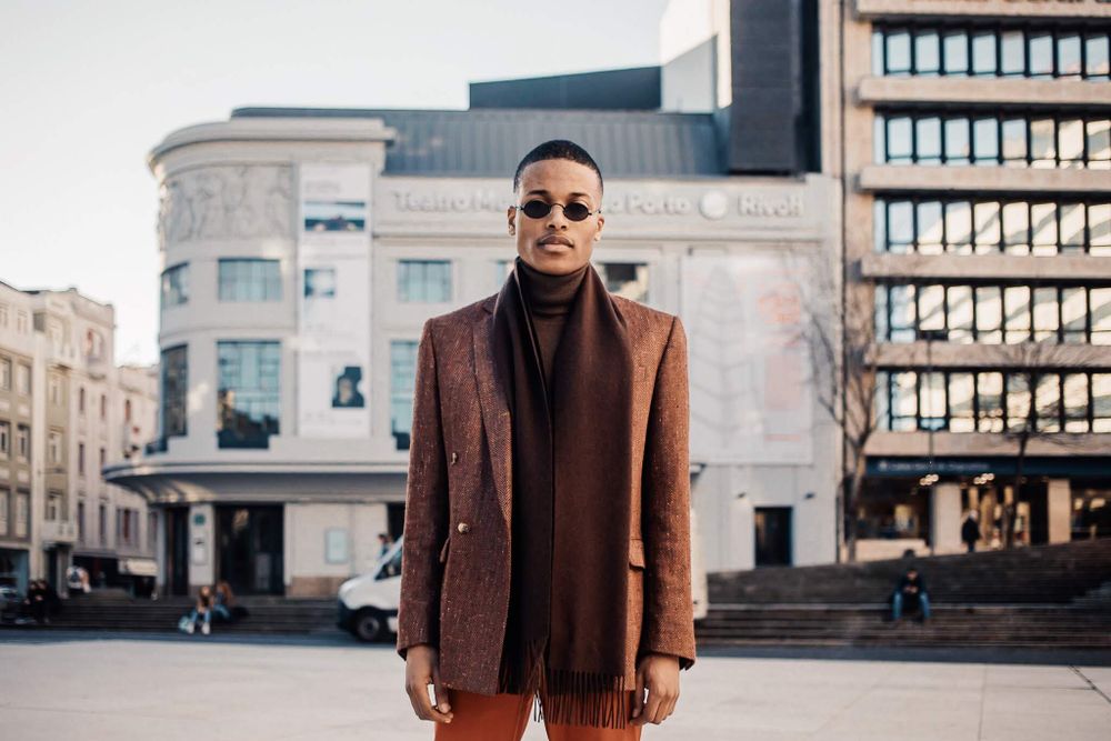 Stylishly dressed person in brown blazer and turtleneck poses on city street against urban architecture backdrop.