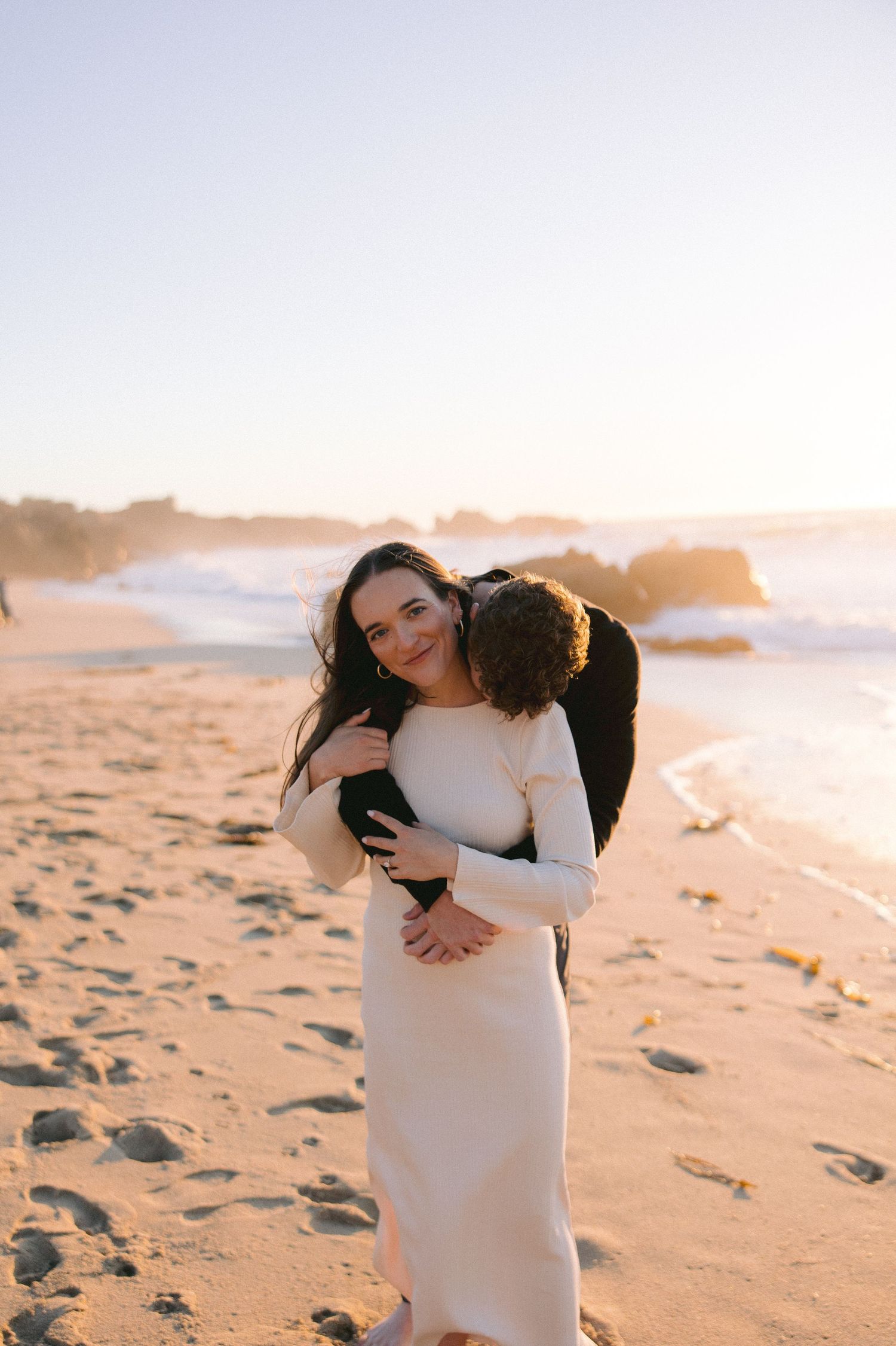 Couple embraces on sandy beach at golden sunset with ocean waves crashing in background.