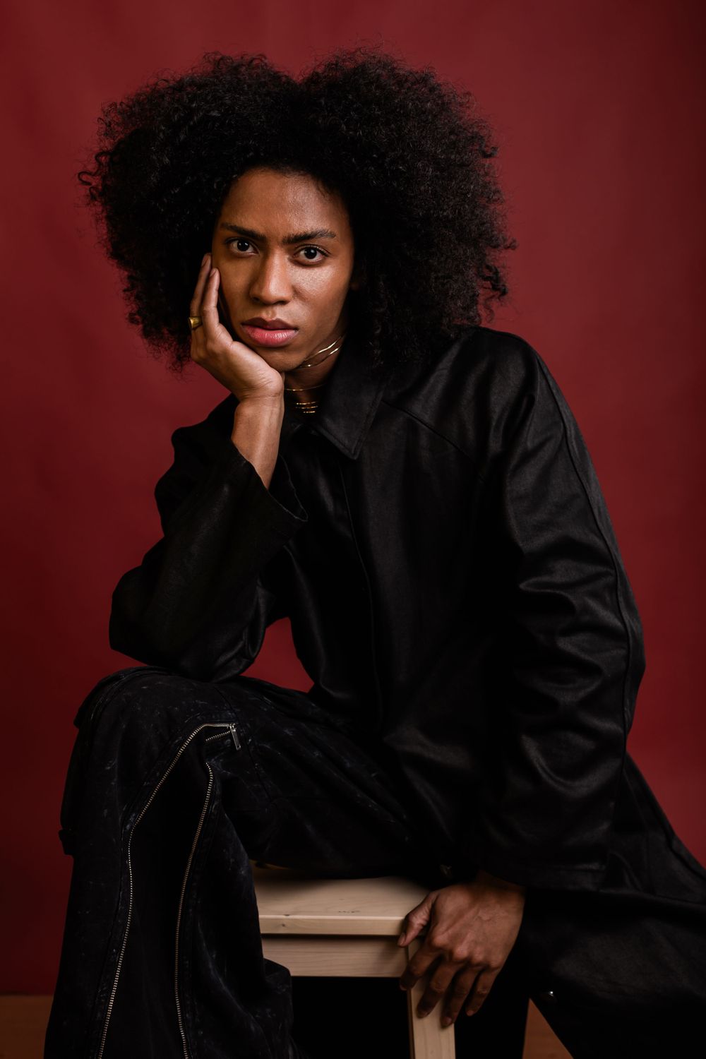 Studio portrait with dark curly hair wearing all black against a deep red background.