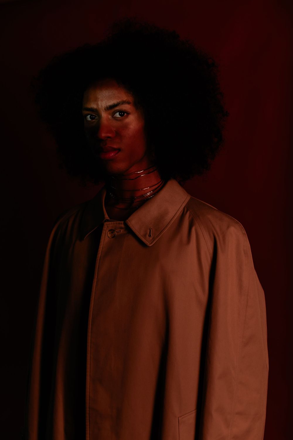 A figure in a tan coat poses against a dark red background with dramatic shadow lighting and natural hair.