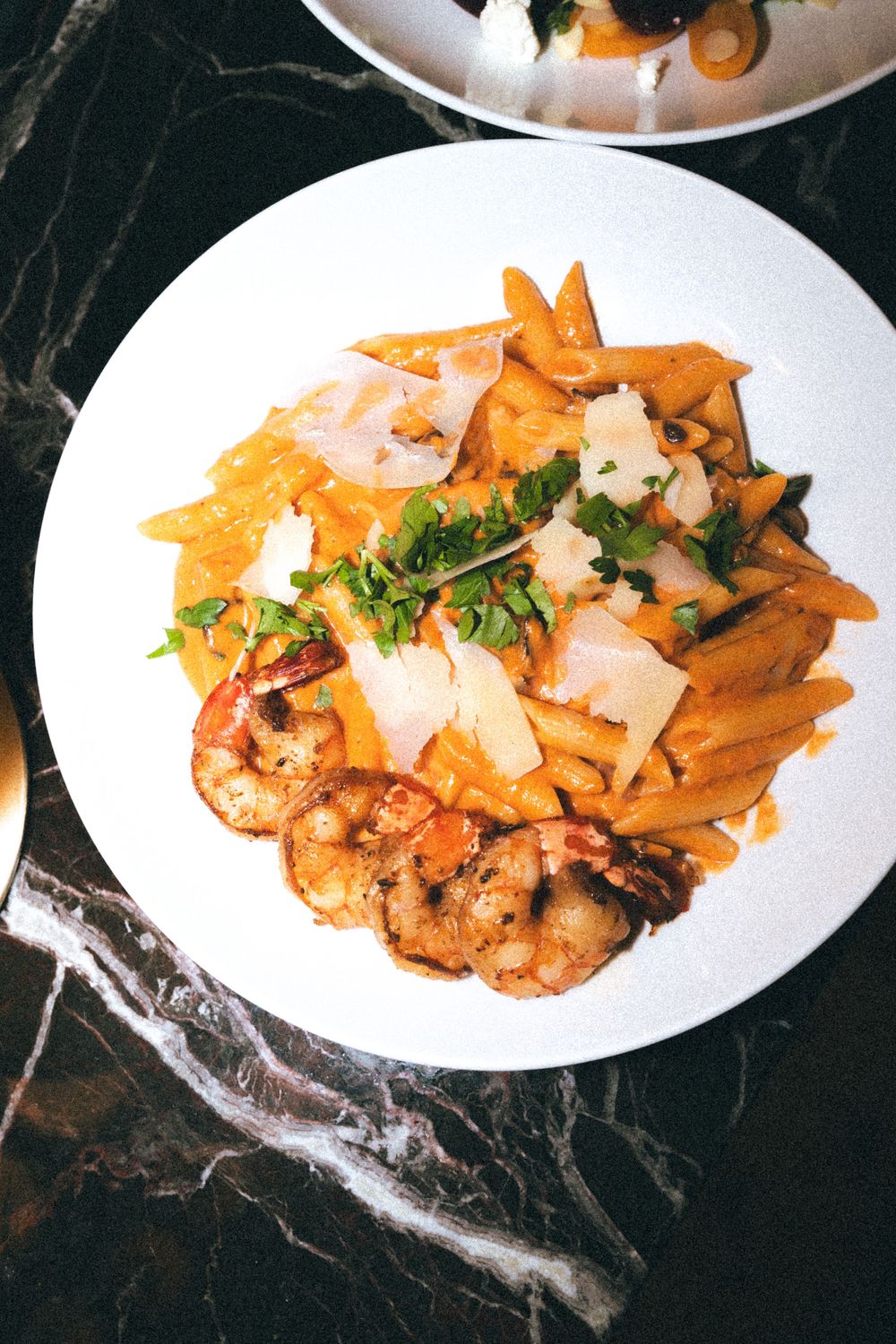 A gourmet pasta dish with shrimp and parmesan shavings served on a white plate atop a dark marble surface.