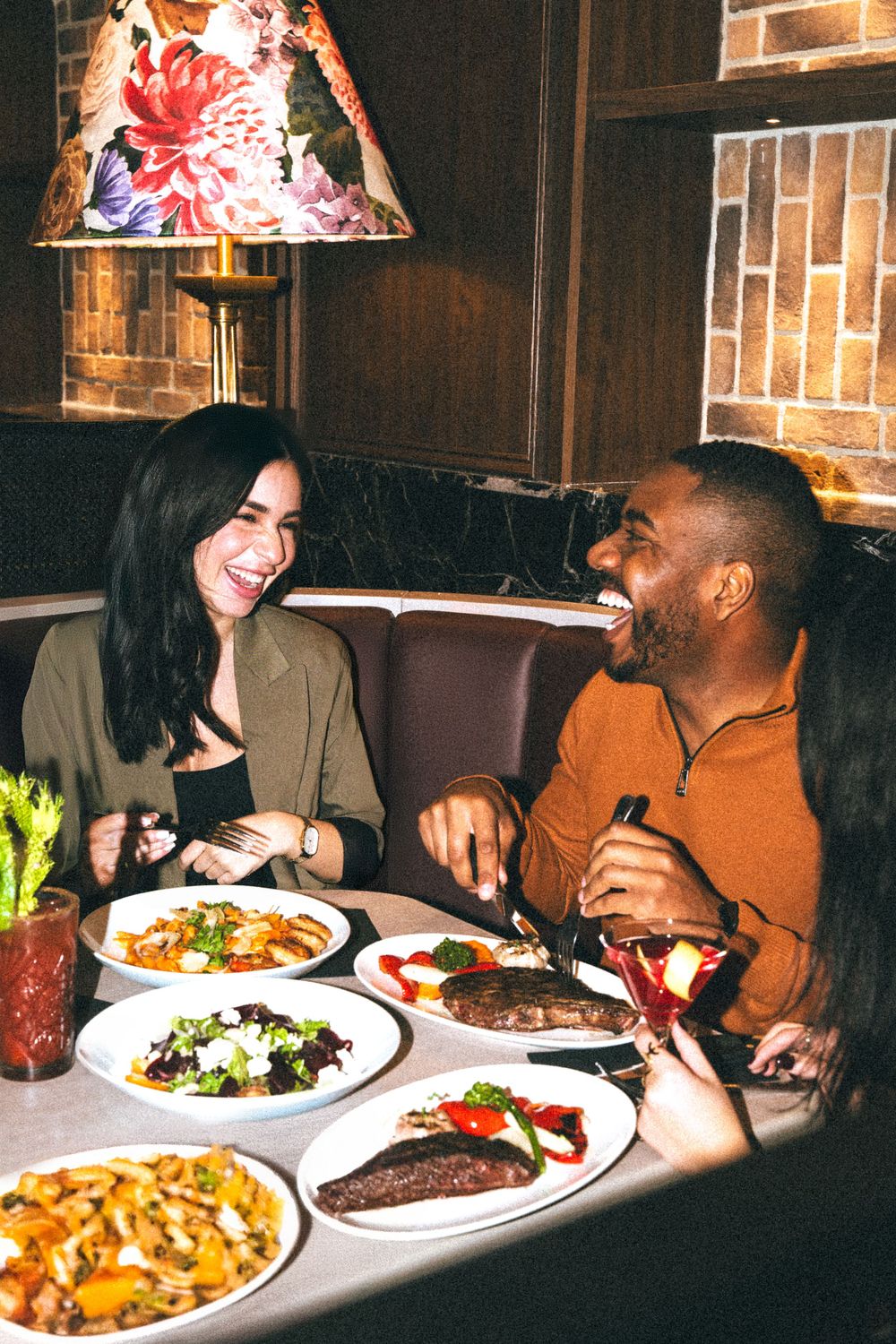 Two diners share laughs while enjoying multiple dishes including steak and salad at a cozy restaurant with brick walls.