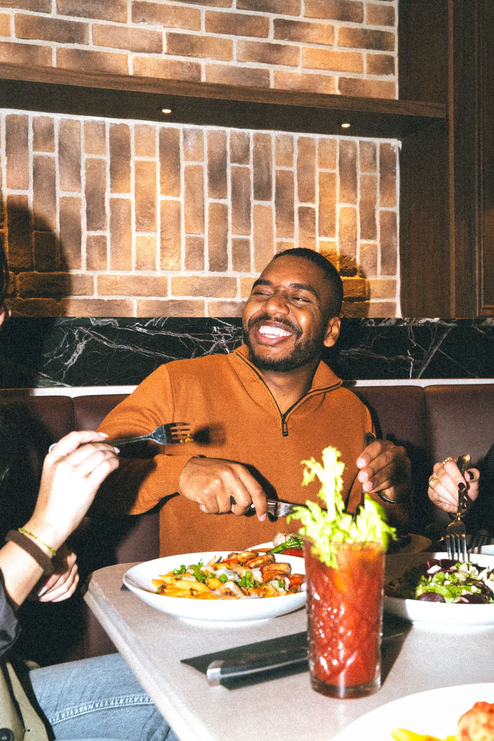 Someone enjoying a meal at a restaurant with a warm smile, wearing an orange sweater, with a Bloody Mary cocktail on the table.