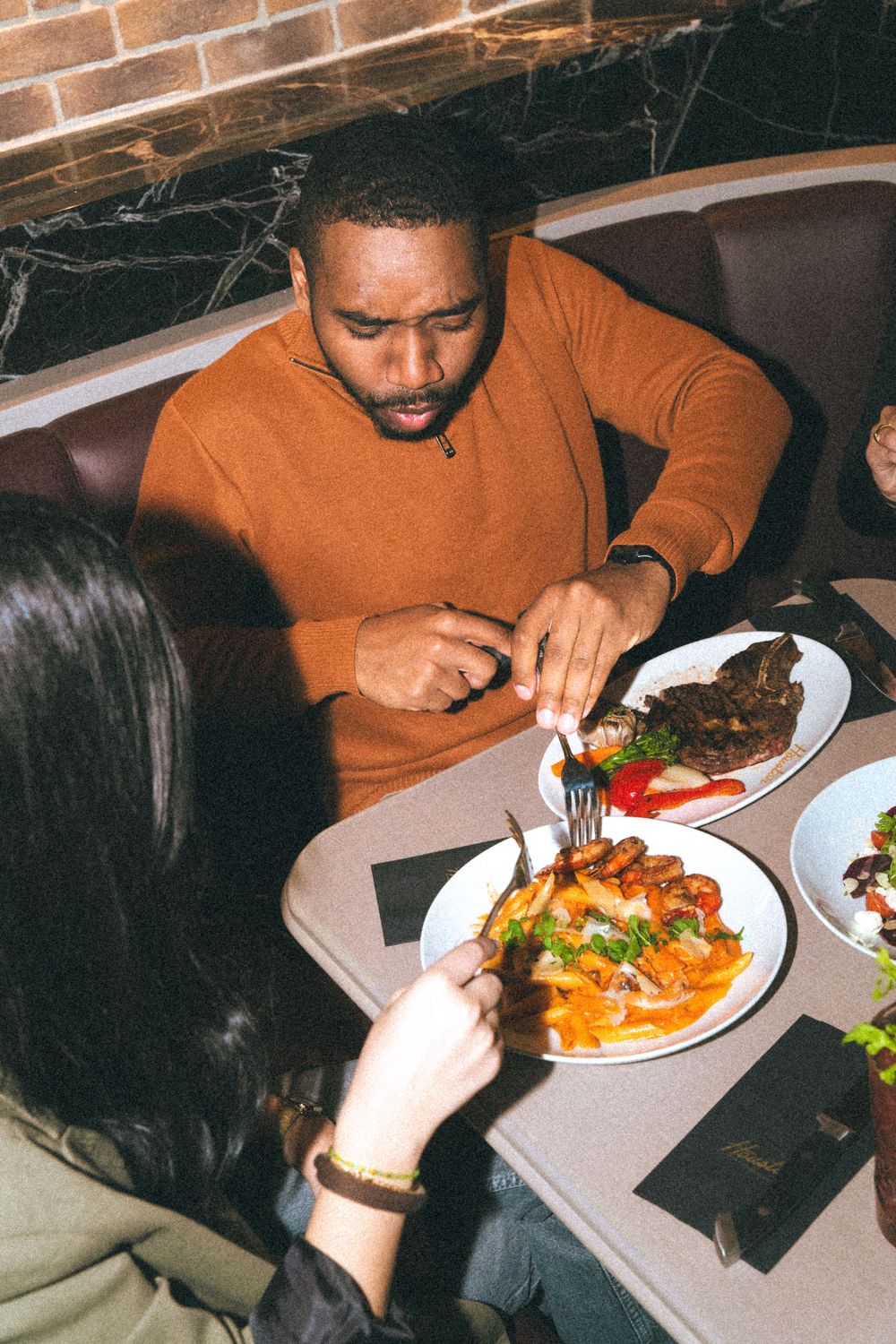 A person in an orange sweater enjoying a meal with colorful dishes at a restaurant with marble table surfaces.
