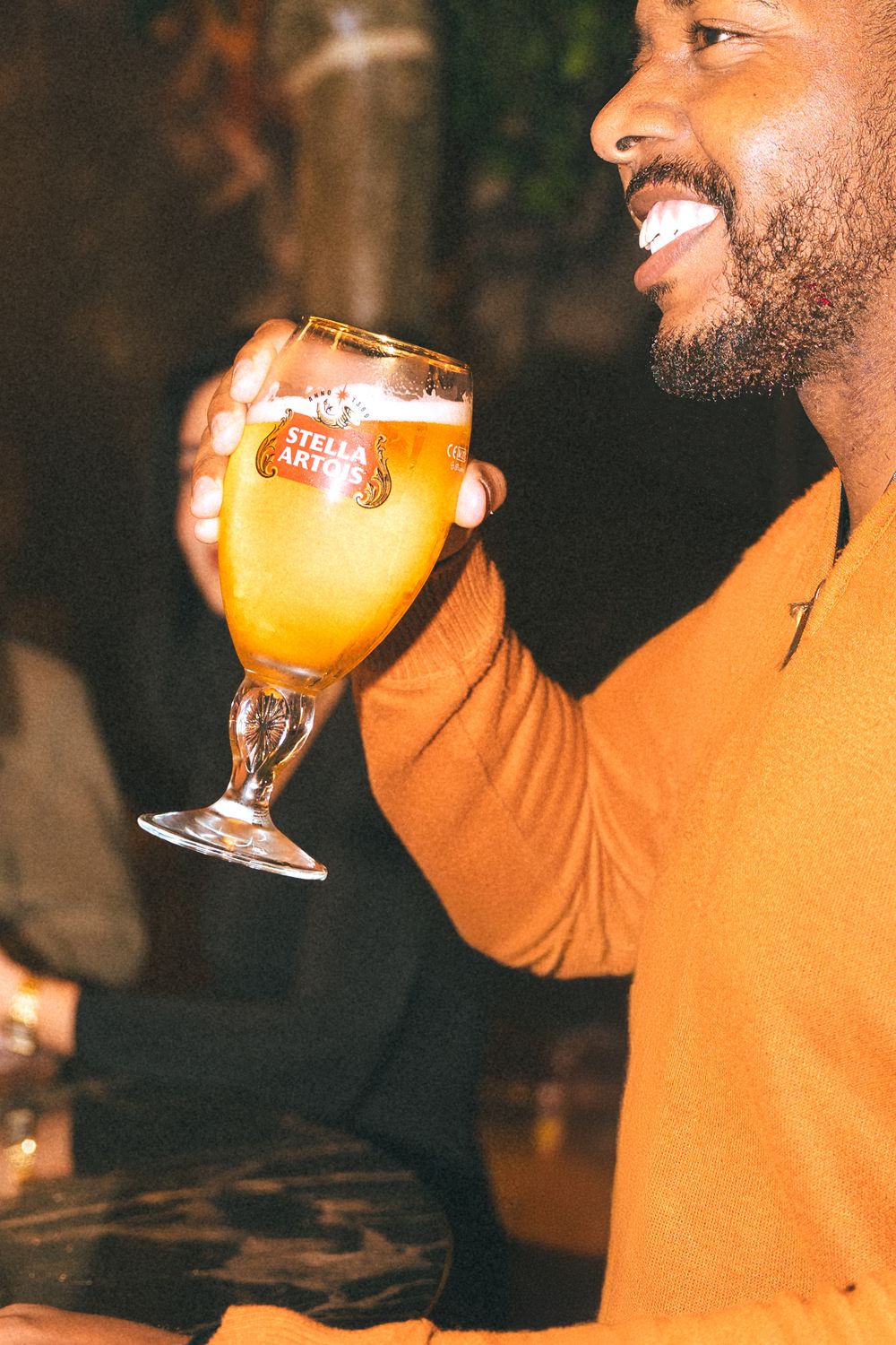 Someone wearing an orange sweater holds up a glass of golden Belgian beer with a happy expression.