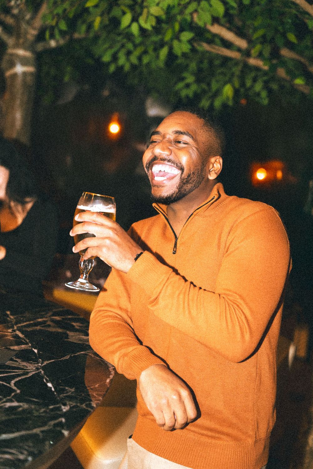A person in an orange sweater laughs joyfully while holding a glass of beer outdoors at night.