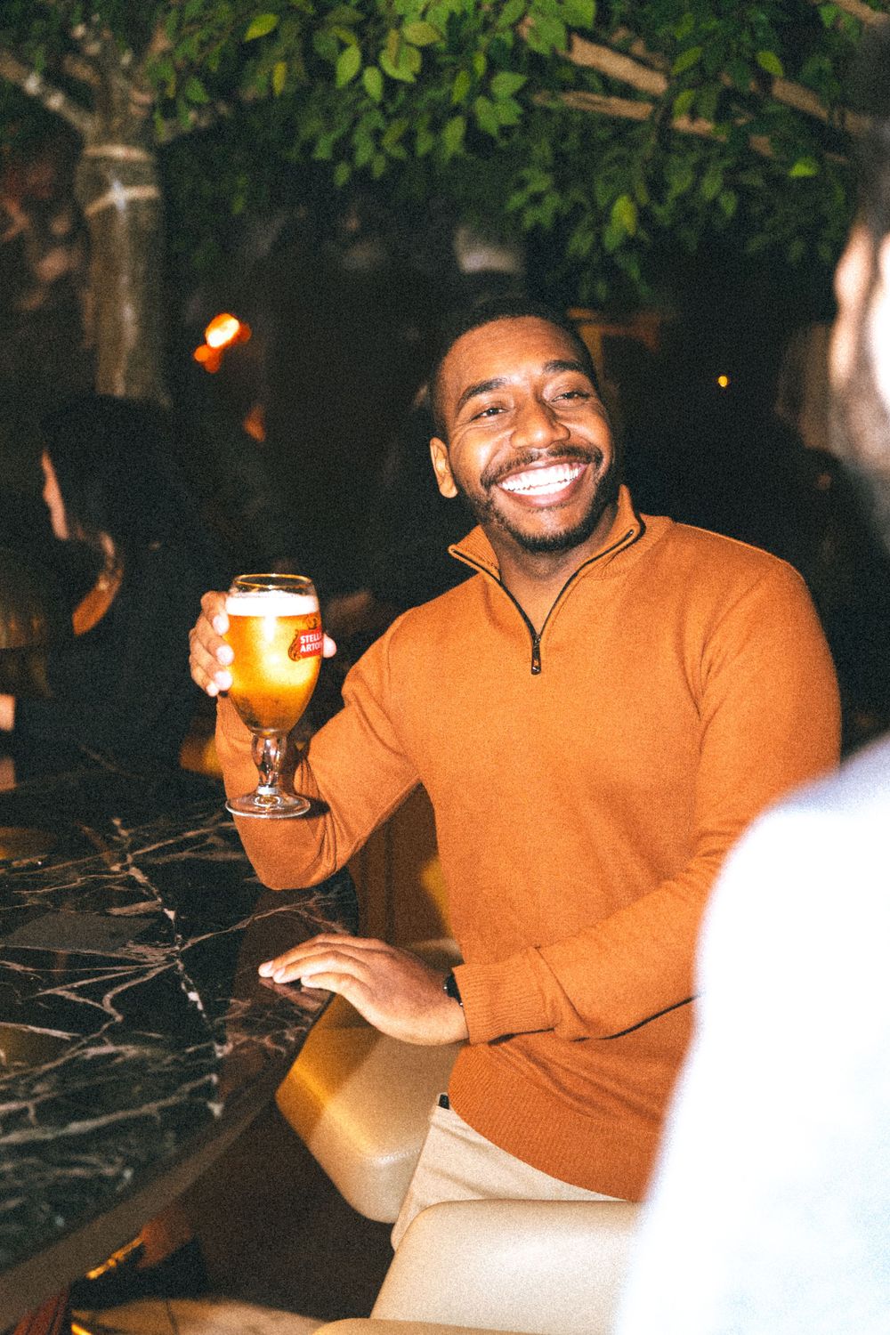 Person in orange sweater smiles while holding a beer glass at outdoor gathering during evening.