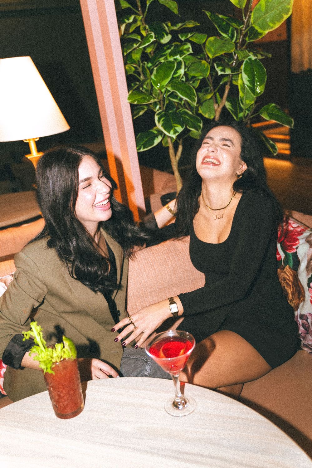 Two friends share a candid moment of laughter at a dimly lit restaurant table with cocktails and indoor plants.
